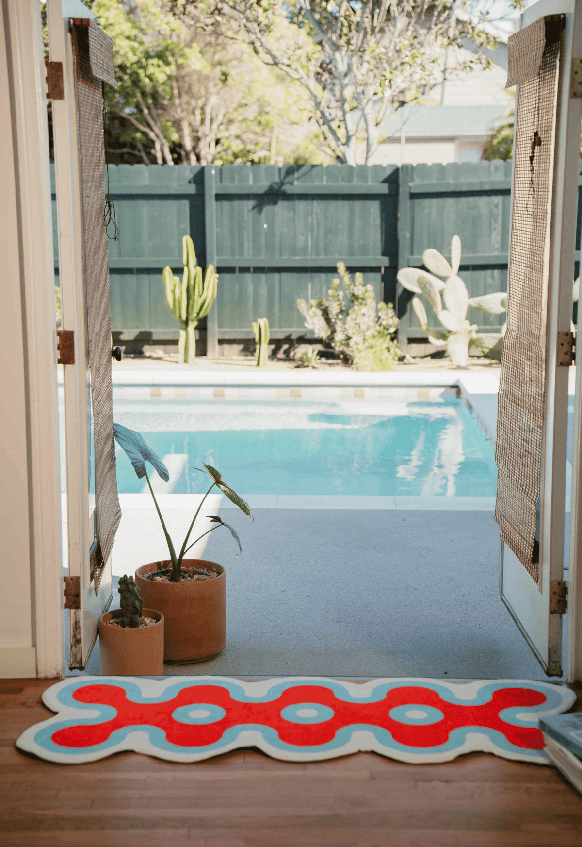Red coral runner rug with aqua trim by open doorway leading to sunny backyard pool with potted plants.