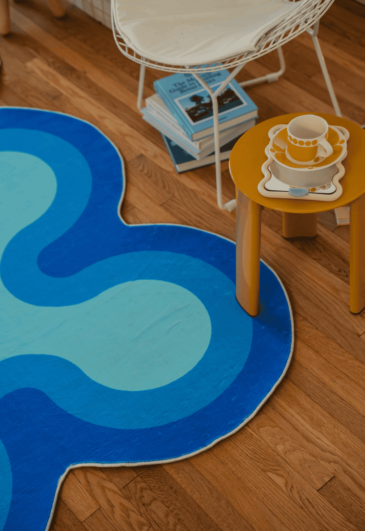 Corner view of blue coral-patterned rug next to a yellow side table with stacked dishes and a white chair.