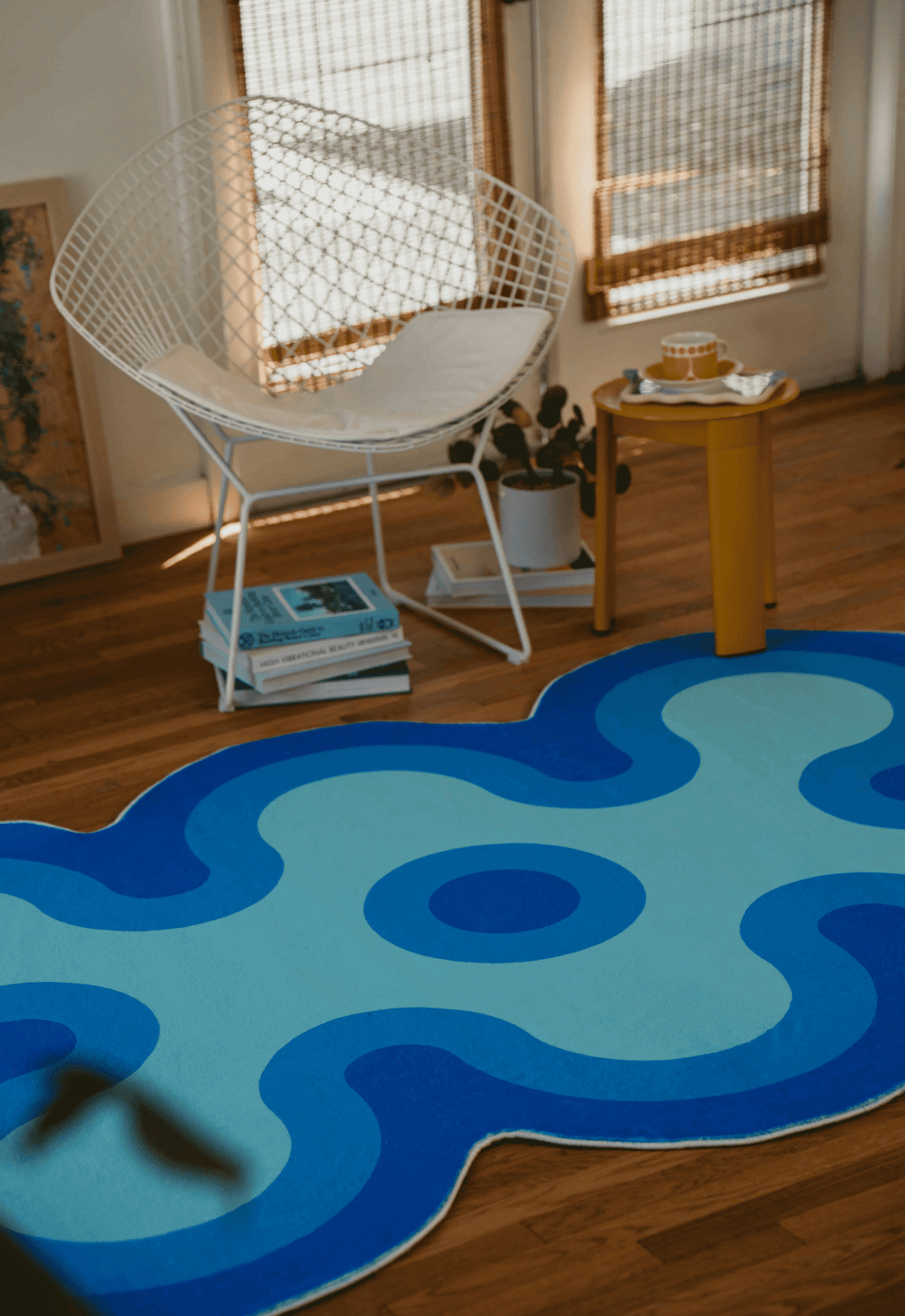 Blue gradient coral-shaped rug in front of a white wire chair, yellow side table, and books stacked on wooden floor.