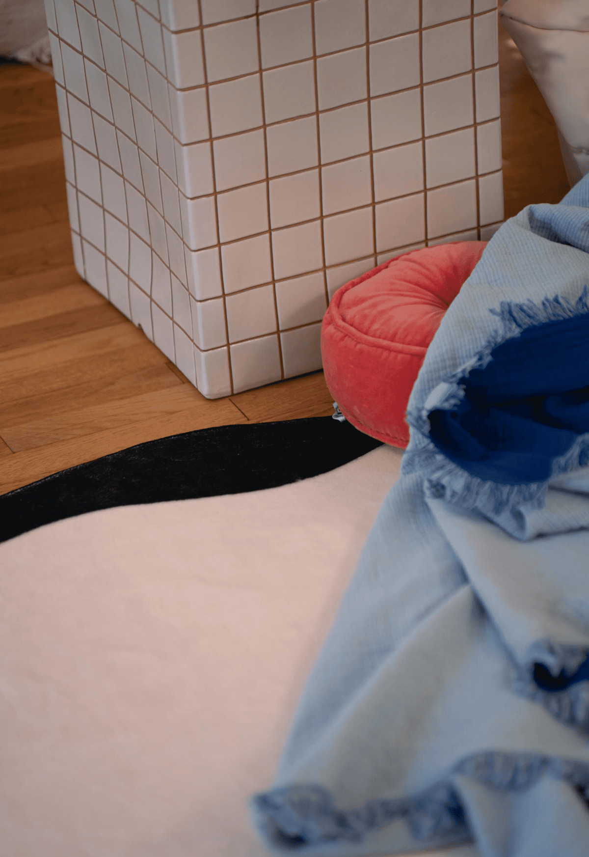 A close-up of the corner detail of the ivory rug with a wavy black border, placed near a white tiled side table and pink cushion on wood floor.