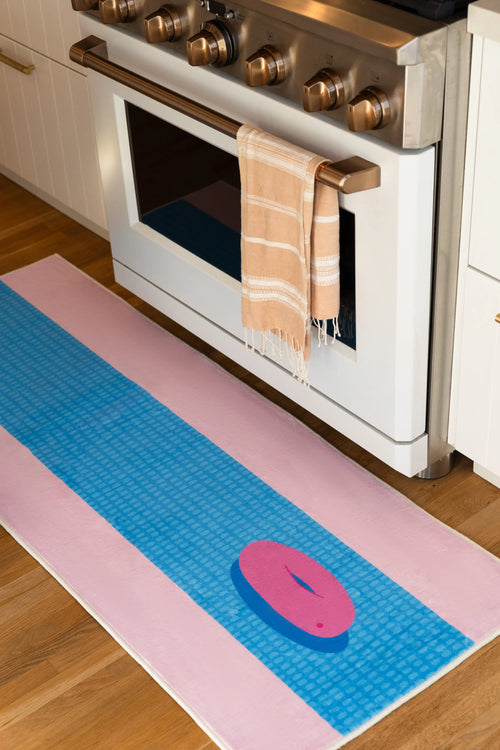 Blue and pink funky pool runner rug beside a white kitchen oven on a light wood floor, with a pink pool float motif