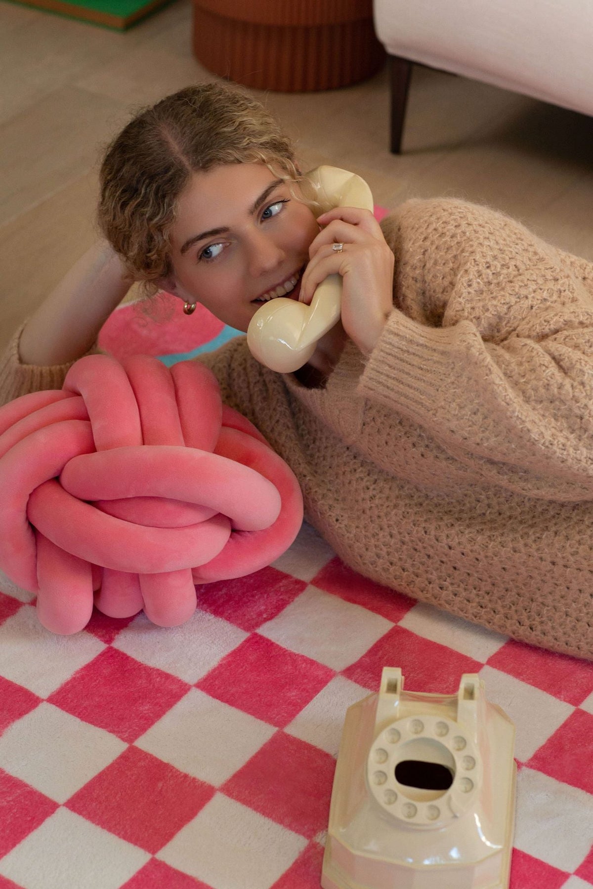 A woman lies on the pink and blue checkered rug, chatting on a retro phone.