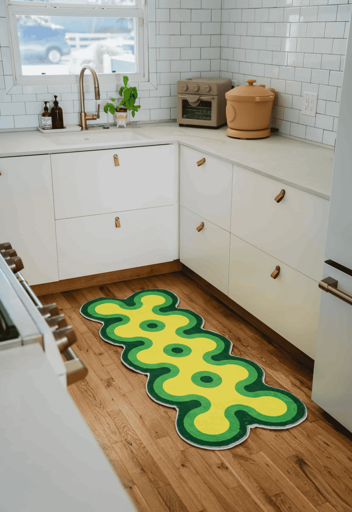 Wavy green and yellow runner rug in kitchen, laid on wood floor near stove and cabinets, with bright daylight from window.