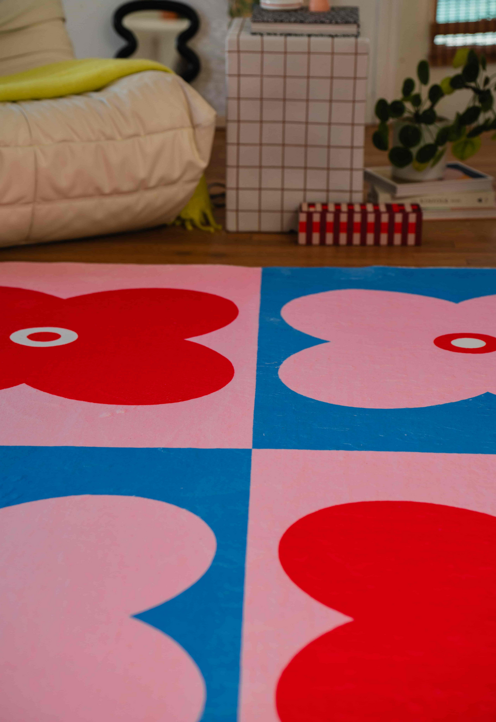 Red and pink flower-patterned rug in front of a cream lounge chair, styled with a grid table and potted plant.