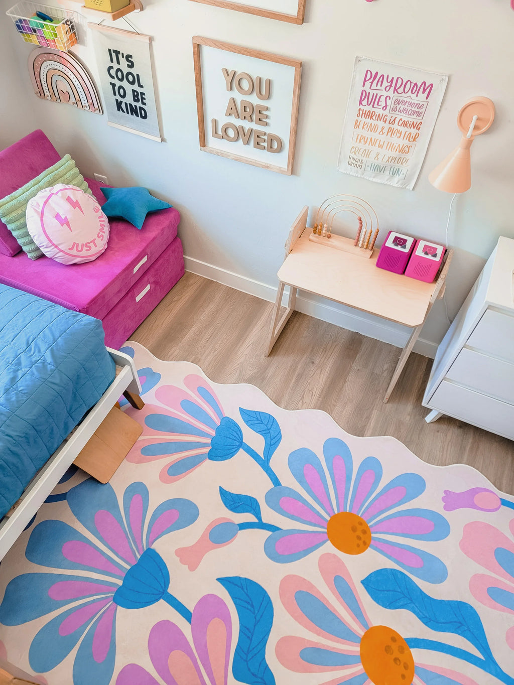 Purple floral wavy rug on pale flooring between a pink bed and wooden desk in a bright bedroom.