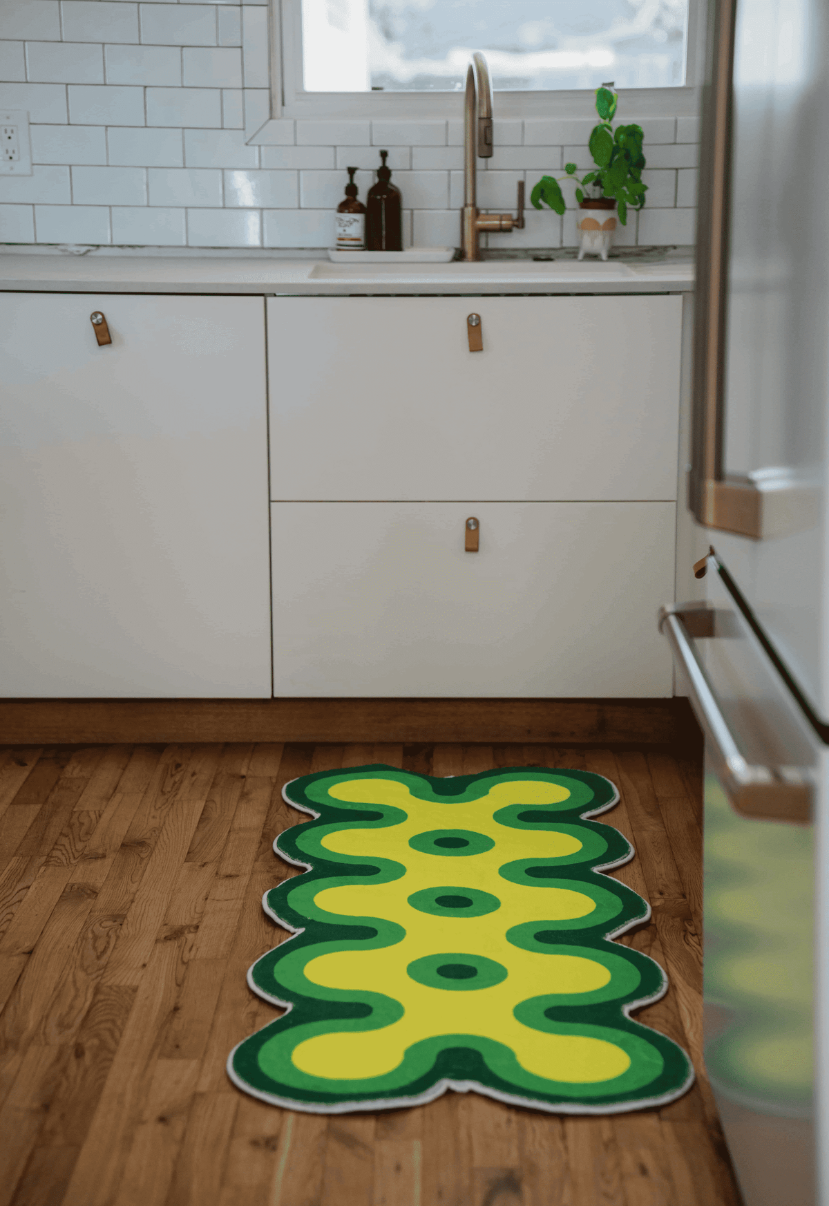 Green and yellow coral runner rug on wood kitchen floor under sink and cabinet, paired with white cabinetry and tile wall.