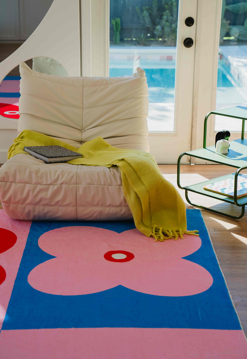 Close-up of a pink and blue clover rug with a cream lounge chair and lime throw in a sunlit room.