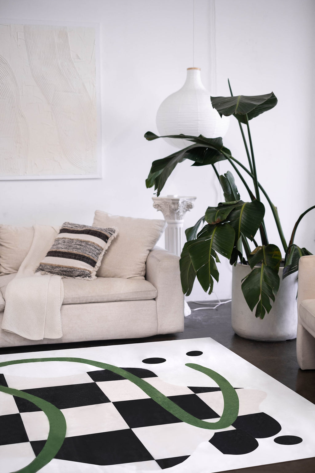 The checkered rug with green curves anchors a minimalist space with a beige sofa, oversized plant, and sculptural pendant light.