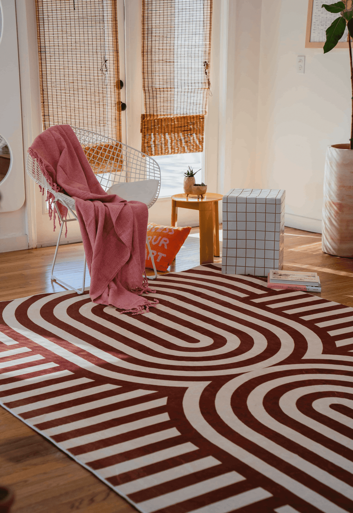 A curvy brown striped rug in a bright room with a wire chair, pink throw, and cube ottoman near wooden steps.