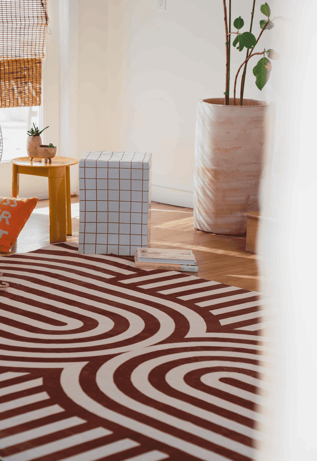 Close-up of the brown striped rug beside a checkered ottoman, books, and tall marbled planter with a fiddle-leaf fig.