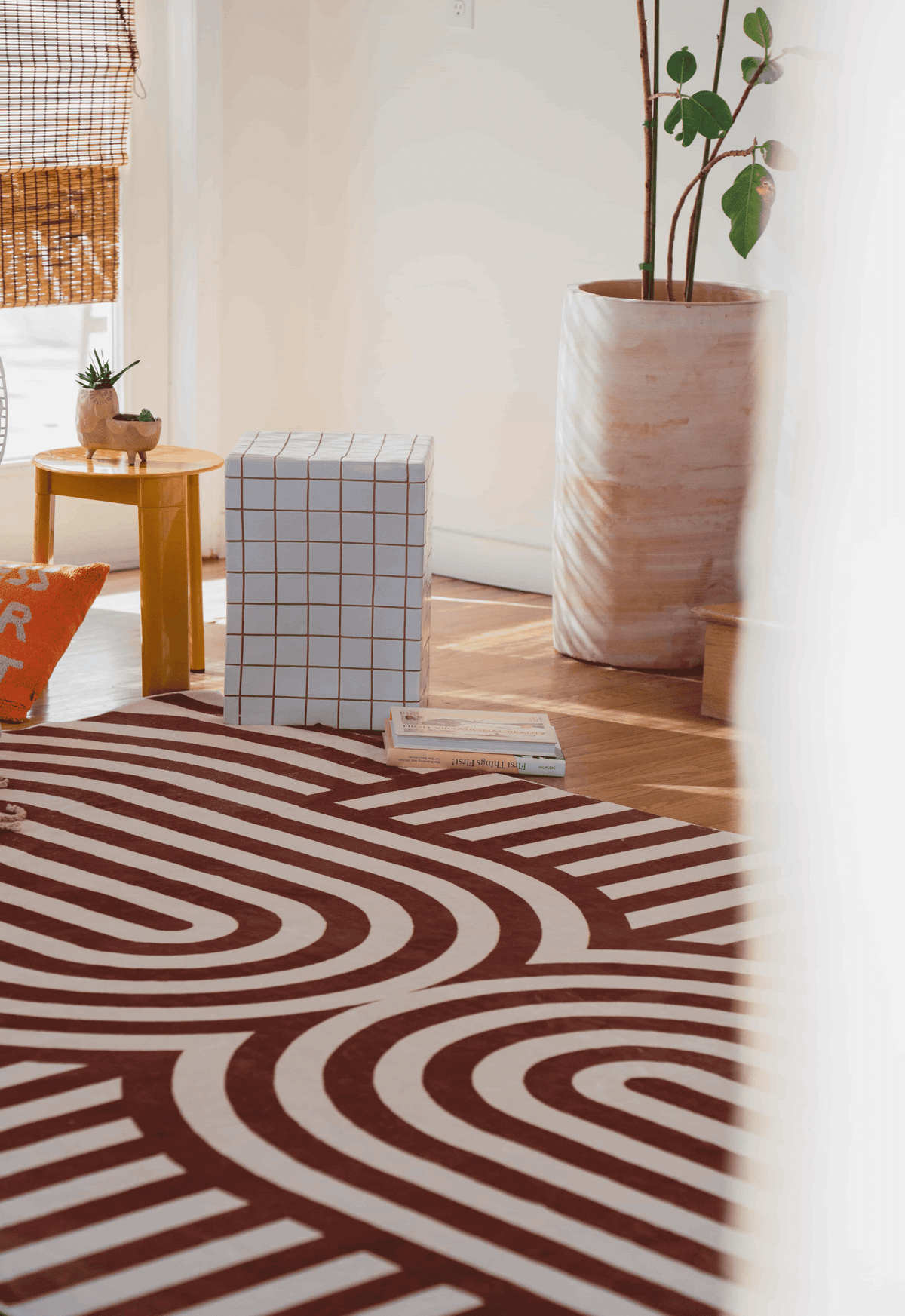 Close-up of the brown striped rug beside a checkered ottoman, books, and tall marbled planter with a fiddle-leaf fig.