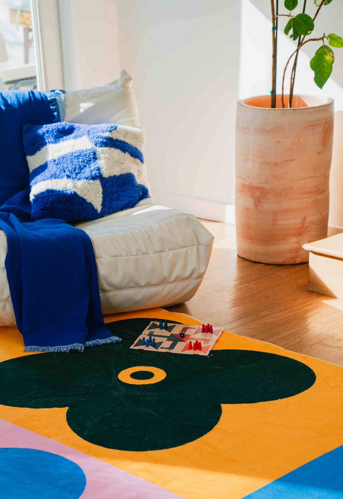 White lounge chair with blue throw sits on green clover rug, next to a large planter in a sunlit room.
