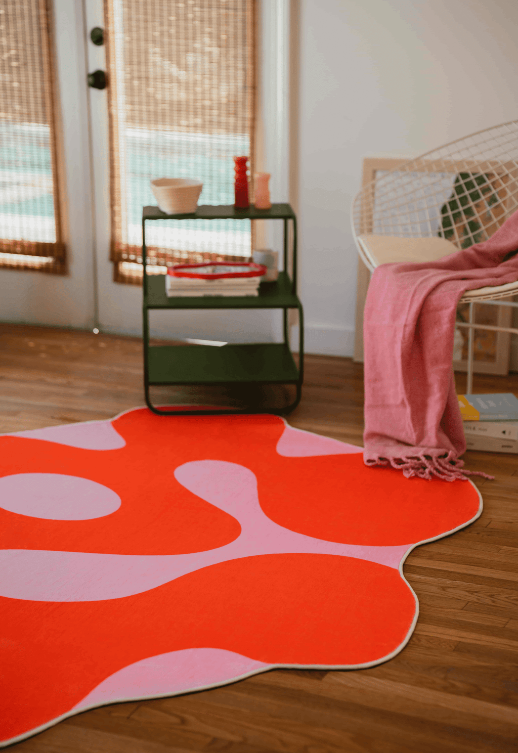 Orange moss rug placed near a wire chair with pink throw and green side table, on natural wood floor by the window.