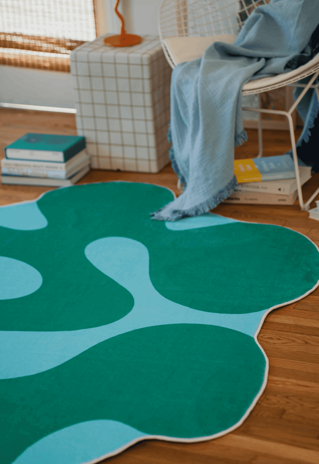 Close-up of green moss rug next to white chair with blue throw and stacked books on a wooden floor.