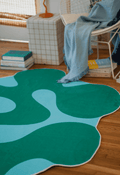 Close-up of green moss rug next to white chair with blue throw and stacked books on a wooden floor.