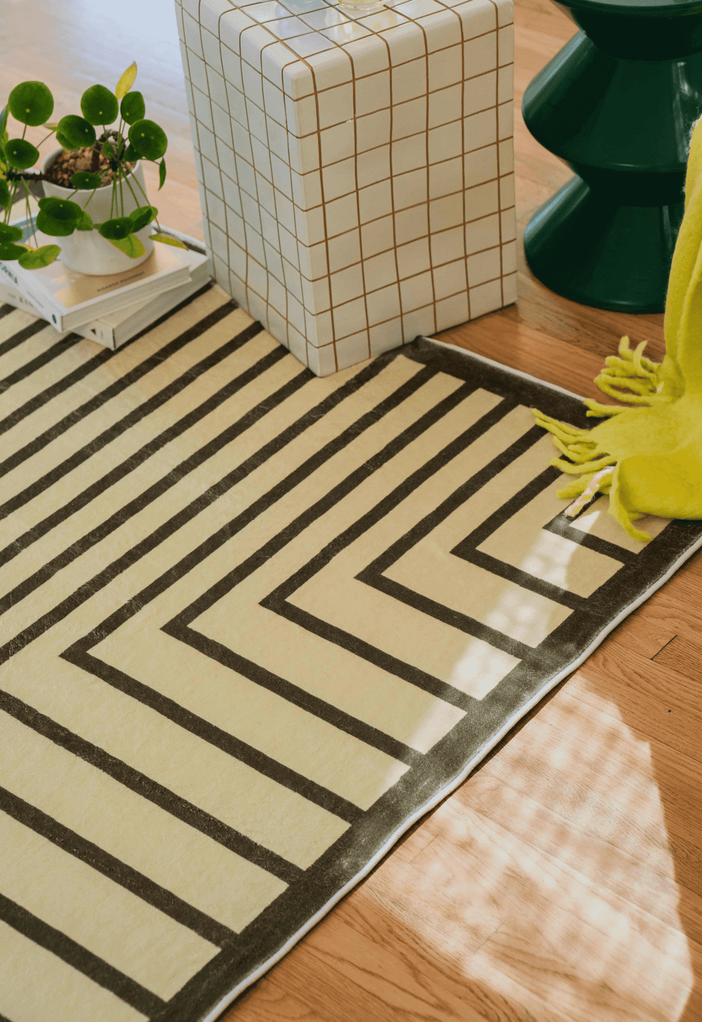 Close-up of olive green rug corner beside grid ottoman and green side table, yellow throw draped casually.