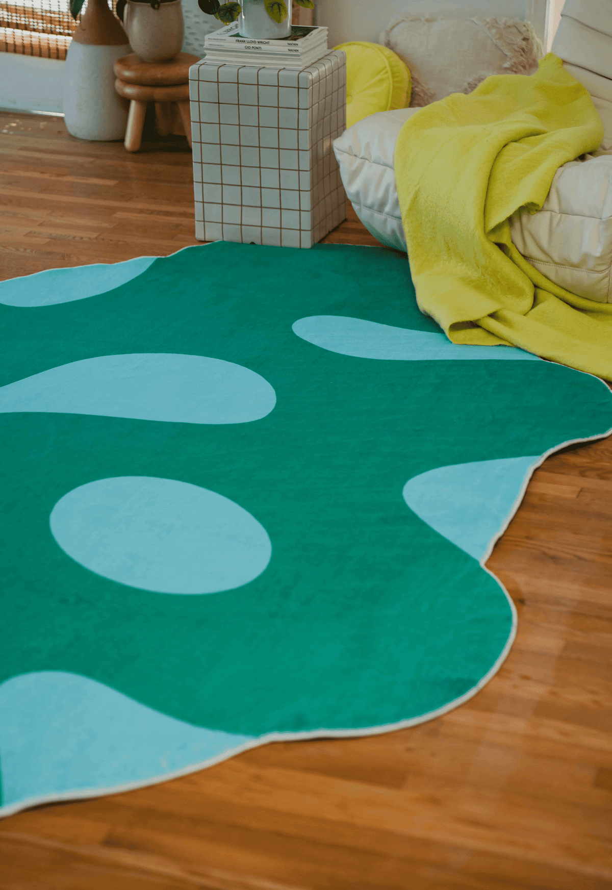 Edge of green moss rug beside couch with yellow throw and checkered side table, on a warm wood floor.