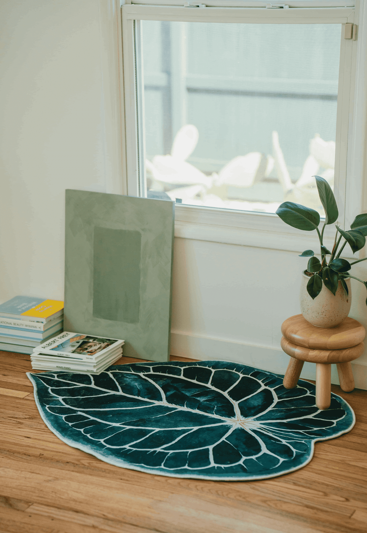 Leaf rug tucked in a bright corner by a window, near a short stool and stacked books on wood floor.
