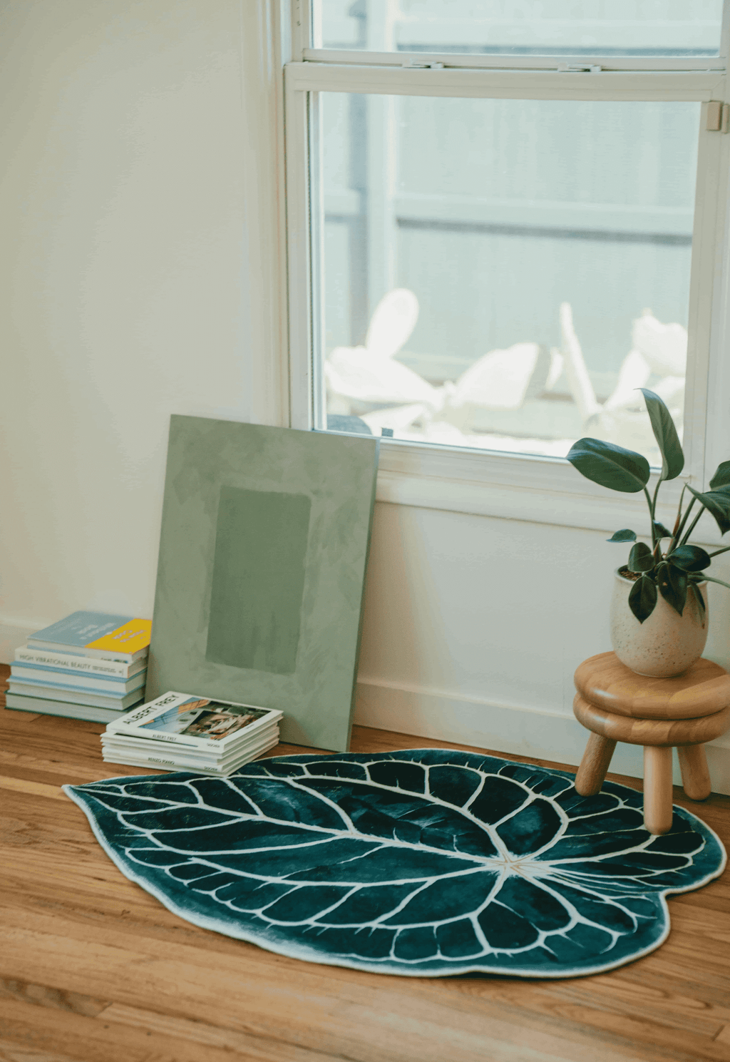 Leaf rug placed under a window beside stacked books and a short stool, in a quiet corner on wood floor.