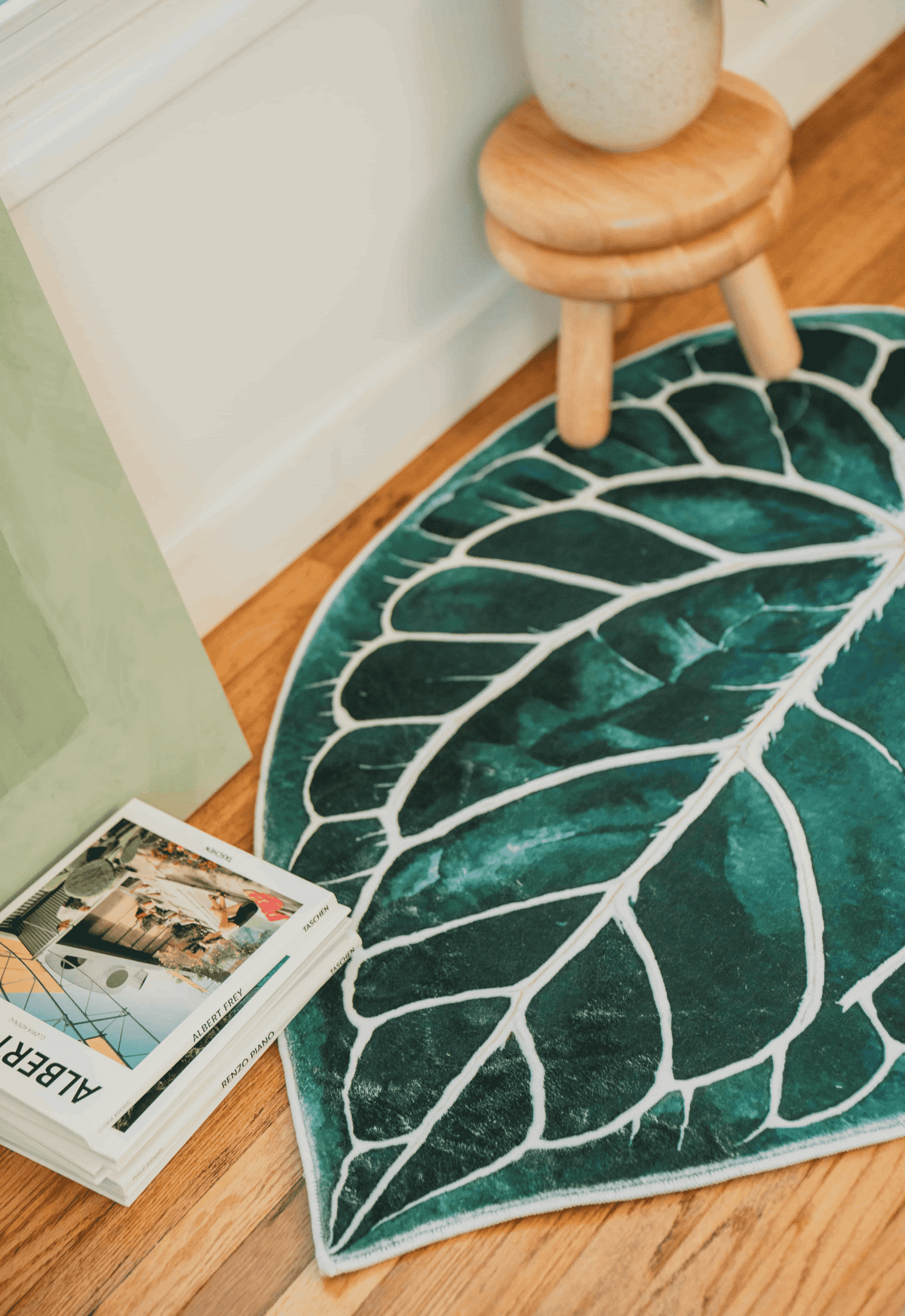 Close-up of leaf rug edge beside stacked magazines and a small stool, resting on smooth wood floor.