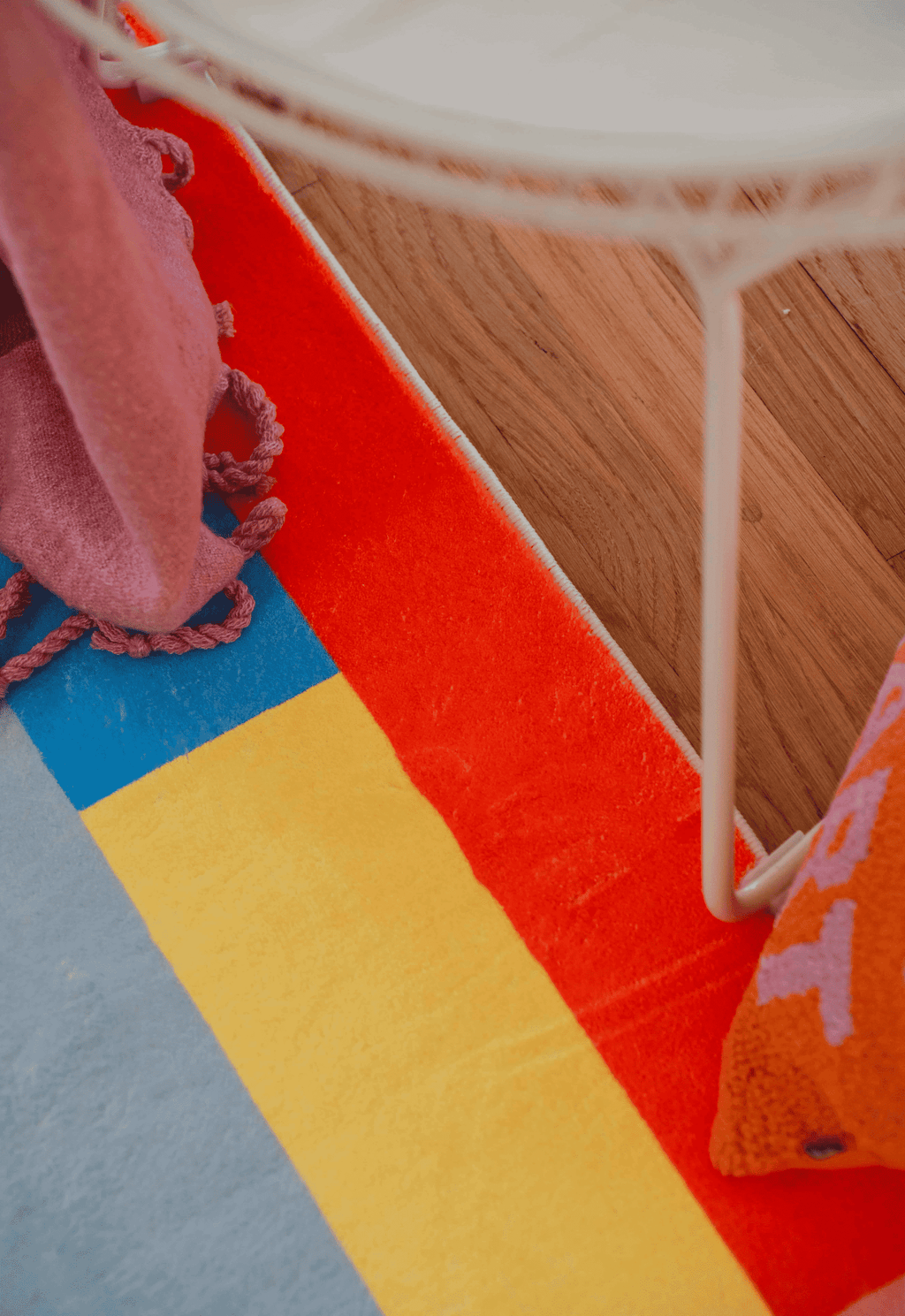 Detail of corner showing red, yellow, and blue geometric borders of the rug, next to a white wire chair and orange text pillow.