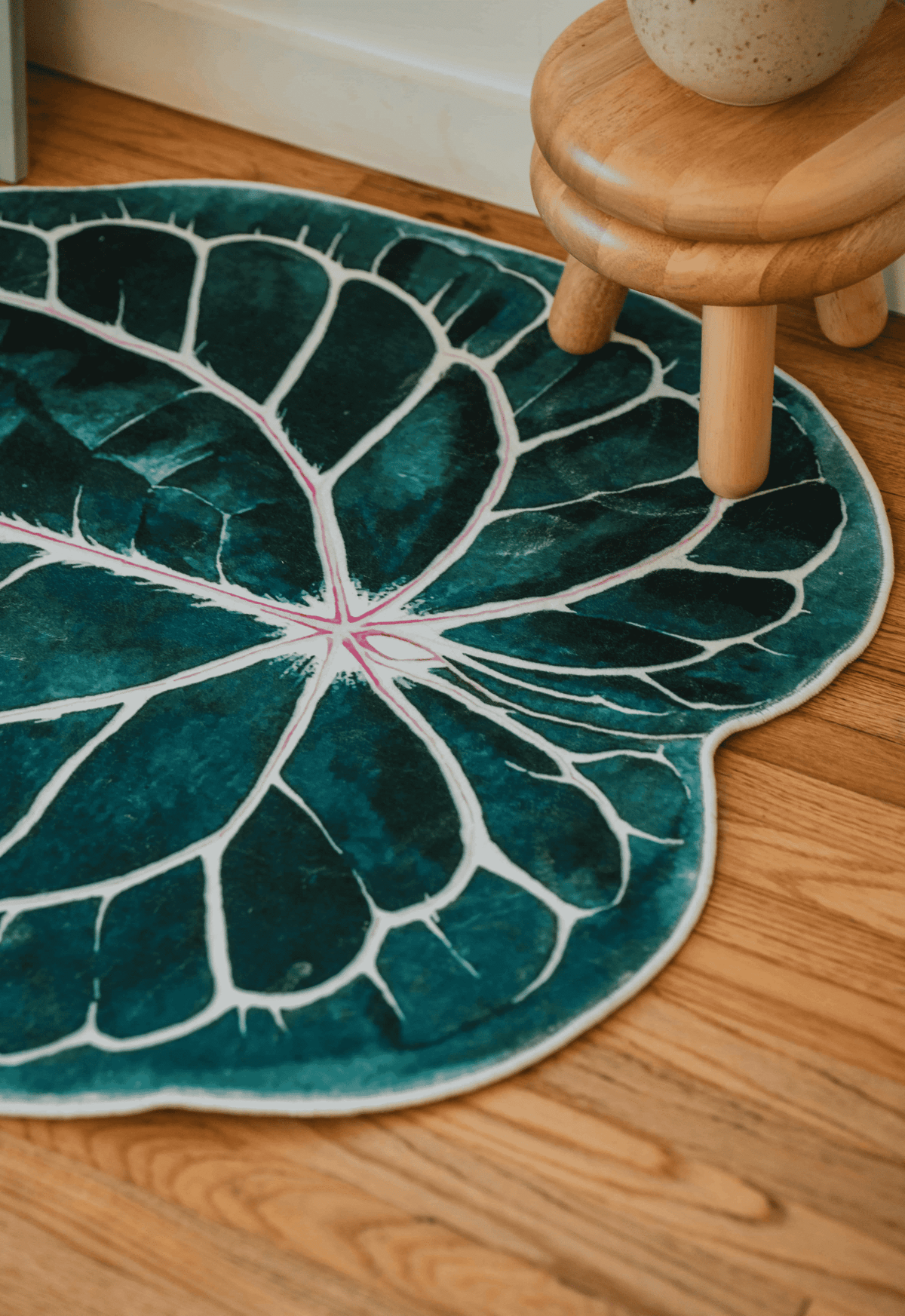 Detail of leaf rug tucked beside a wooden stool and white wall, on smooth wood floor with warm tones.