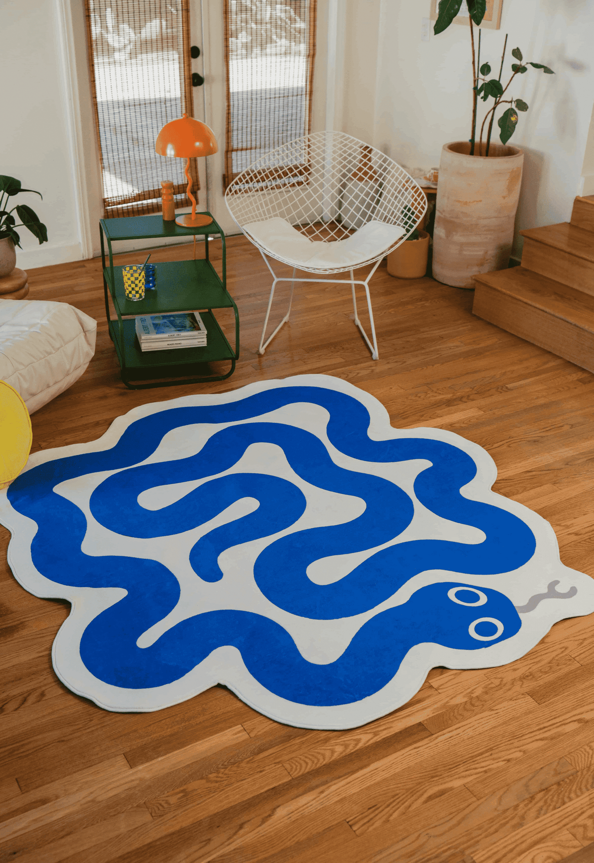 Blue snake rug with white background, placed in a sunny interior with a white wire chair and potted plant nearby.