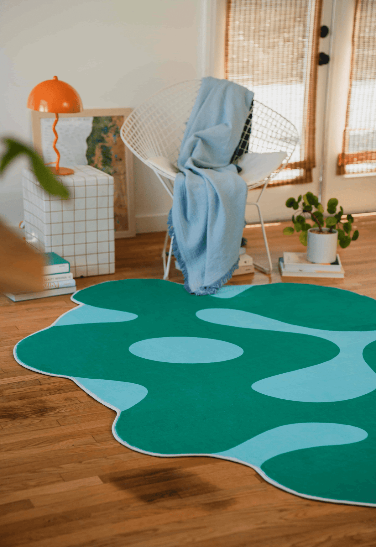 Corner setup with the moss rug under a white chair draped with a blue blanket, orange lamp and indoor plant nearby.