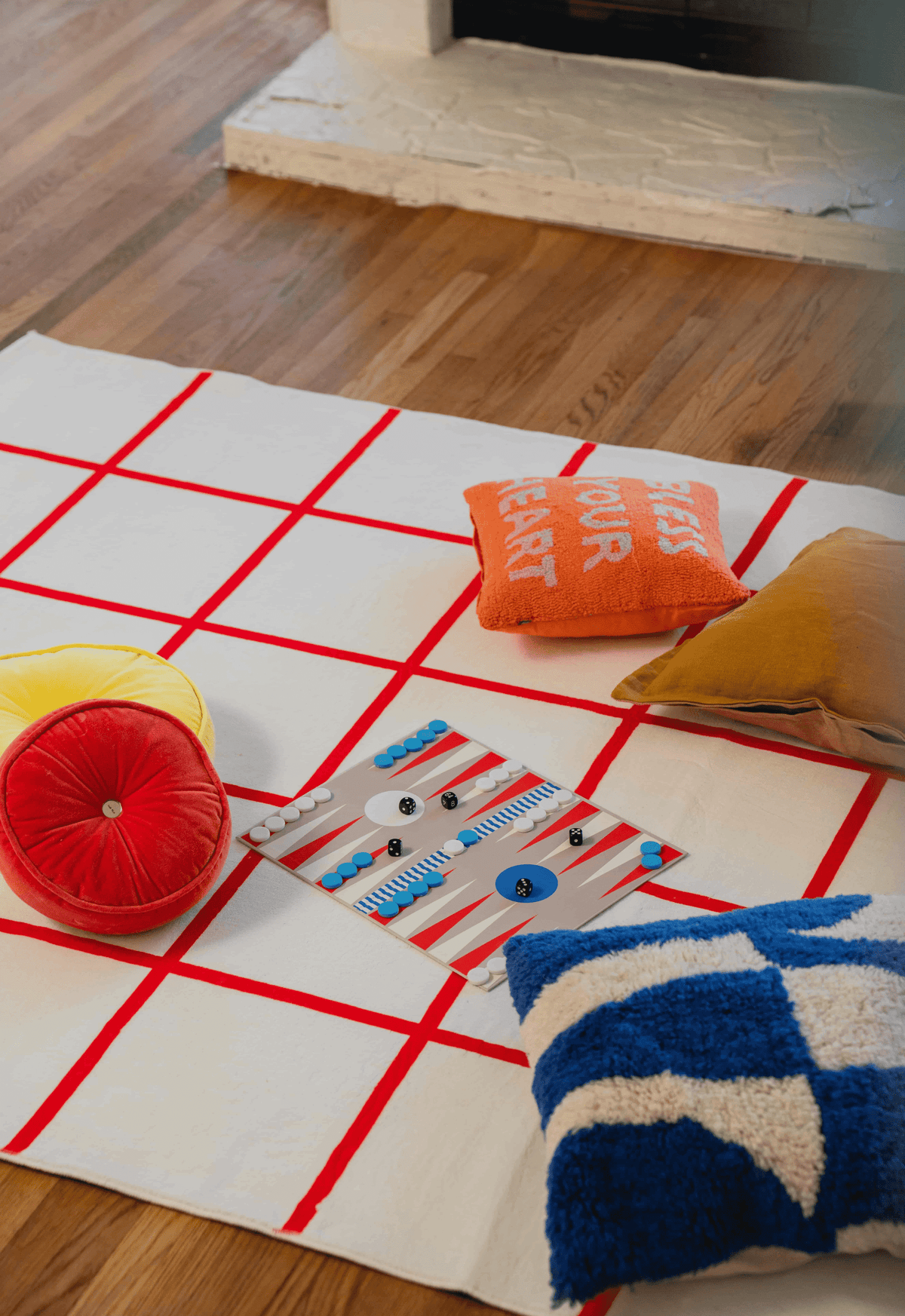 Red and white grid rug as playful floor setup with board game, orange pillow, and velvet citrus cushion near fireplace