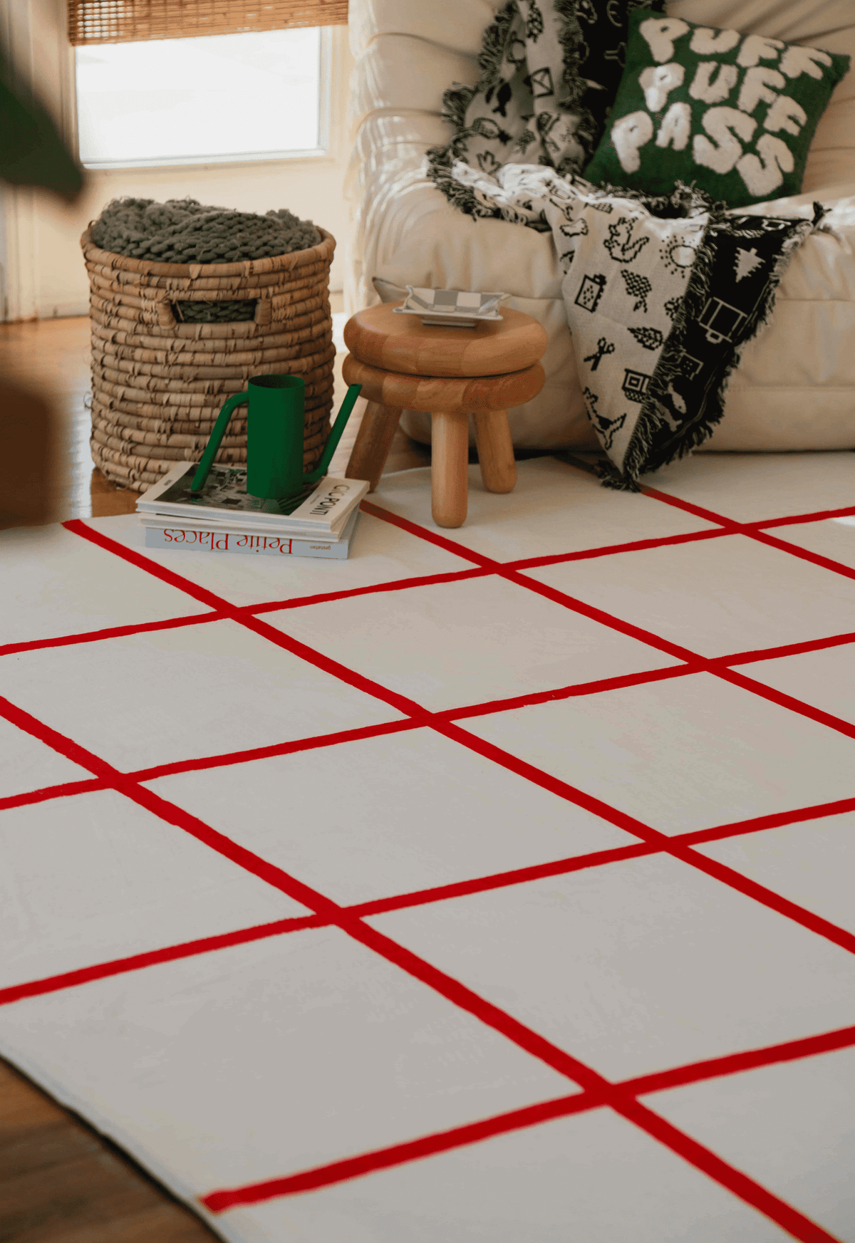 Red and white grid rug under reading chair with patterned pillow, wooden stool, and greenery in relaxed living space.