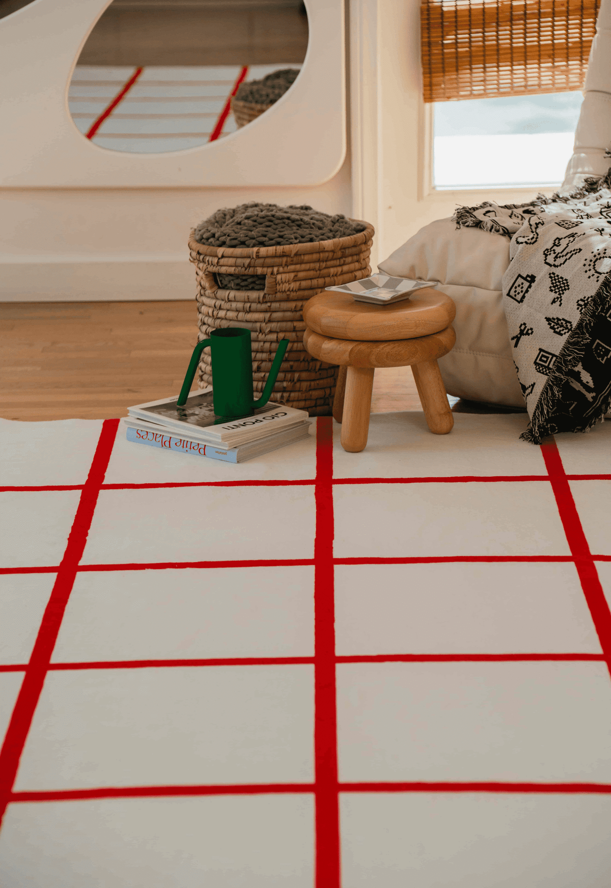 Red grid rug beside stool and storage basket, reflected in wall mirror, creating a cozy corner setup.