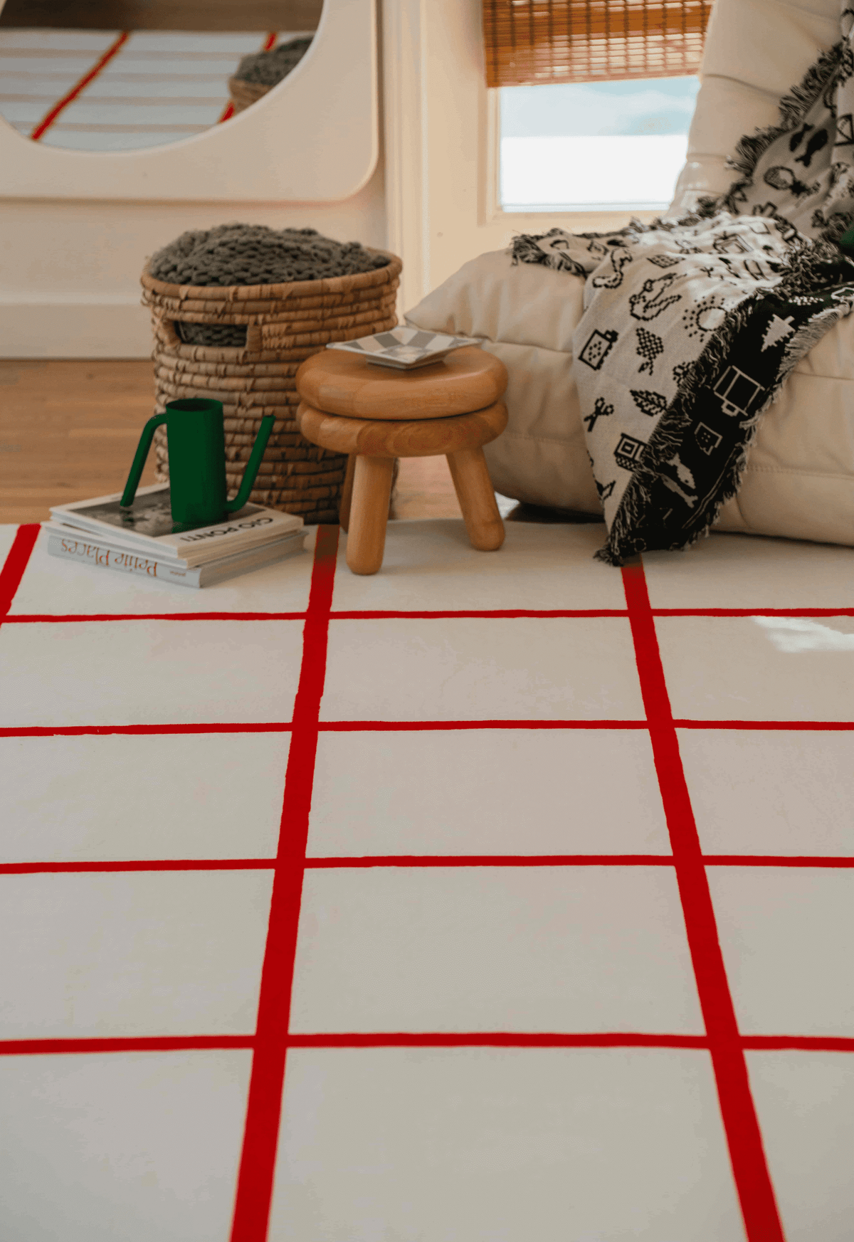 Red and white grid rug in cozy reading nook with cream lounge chair, stacked books, and wooden stool on wood floor.