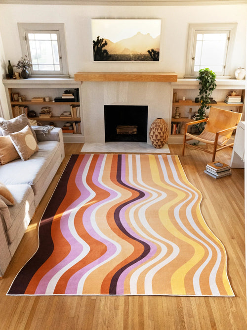 Brown Abstract Striped Rocks Area Rug by Zoe Wodarz in a sunlit living room with a fireplace, shelving, and a wood chair