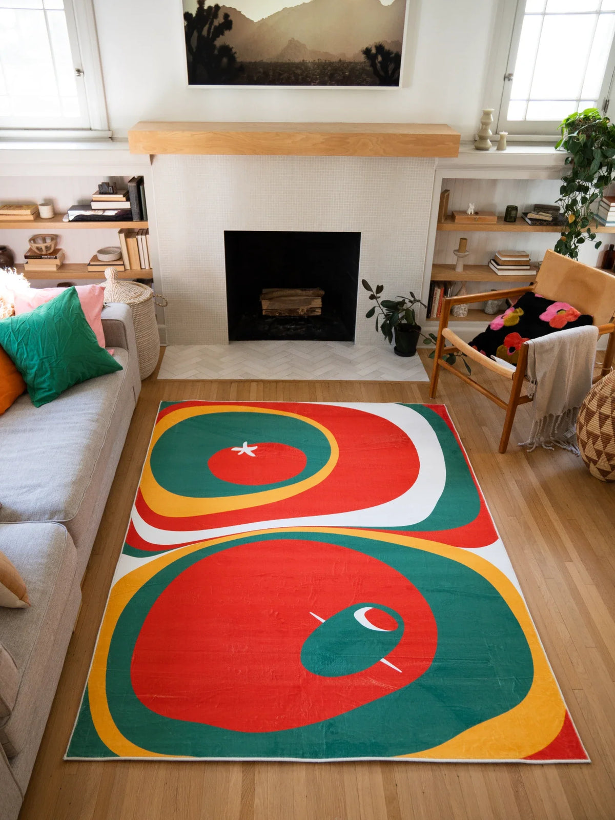 Tomato Twist Abstract Area Rug by Zoe Wodarz in olive and red, styled in a bright living room with a fireplace