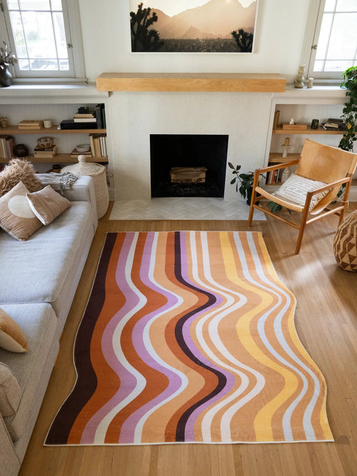 Flowing striped rug framed between a sofa and fireplace, with layered brown, peach, cream, and lilac bands