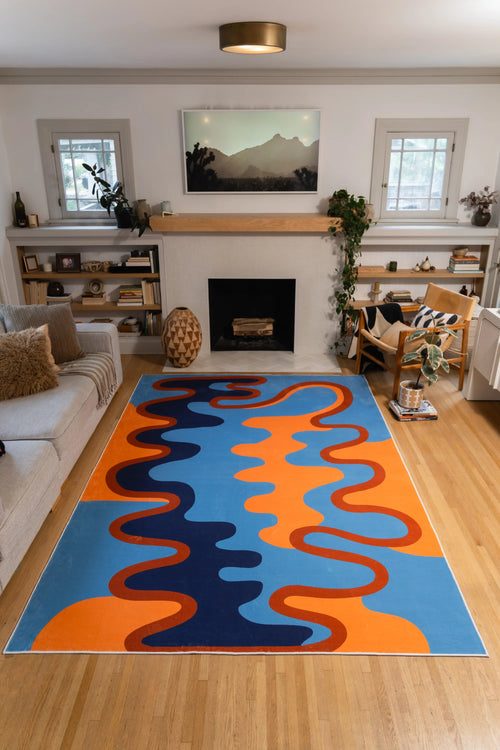The 7' × 10' Blue and Orange Abstract Wave Rug by Ariana Berman stretches between a gray sofa and wooden chair toward the fireplace.