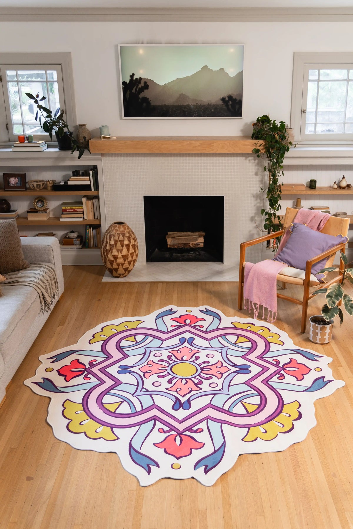 A large, round area rug with a colorful mandala pattern is displayed on a light wood floor in a modern living room setting with a fireplace, chair, and shelves.
