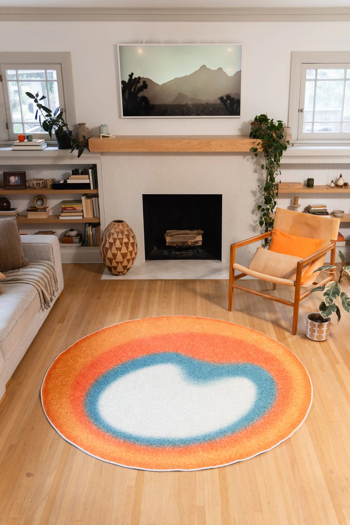 6' × 6' Orange and Teal Abstract Halo Circle Rug by Juan Marquez with an off-white center on a wood floor in front of a fireplace.