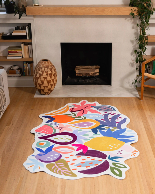 A colorful, fruit-shaped area rug with a white border is displayed on a light wood floor in a cozy living room setting with a fireplace in the background.
