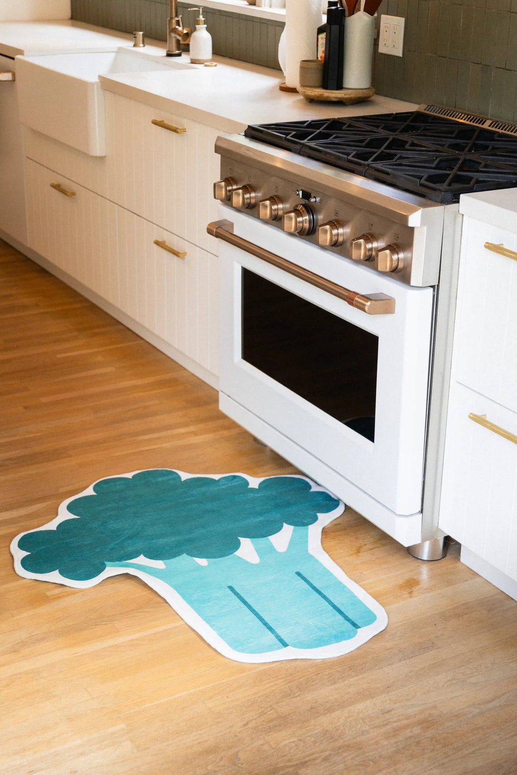 Broccoli-shaped rug with a dark green crown and teal stem on a wood floor beside a white oven and cabinets.