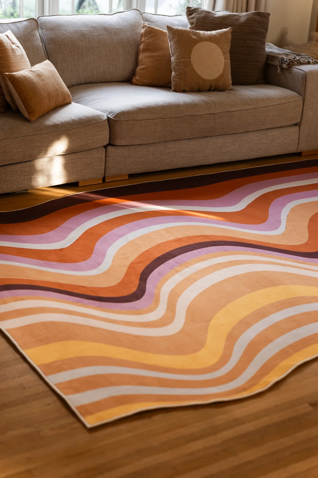 Close view under a beige sofa showing the rug's curved rock like bands in brown, rust, blush, and cream