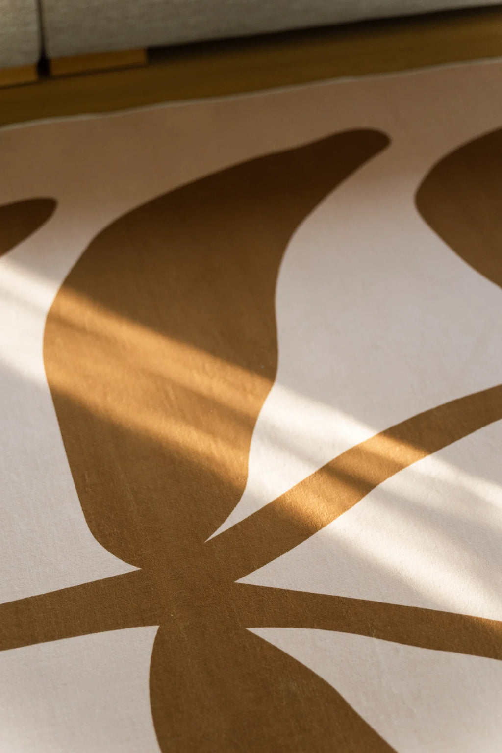 Abstract botanical rug detail with sweeping brown leaf forms crossing a warm beige ground beneath a sofa edge