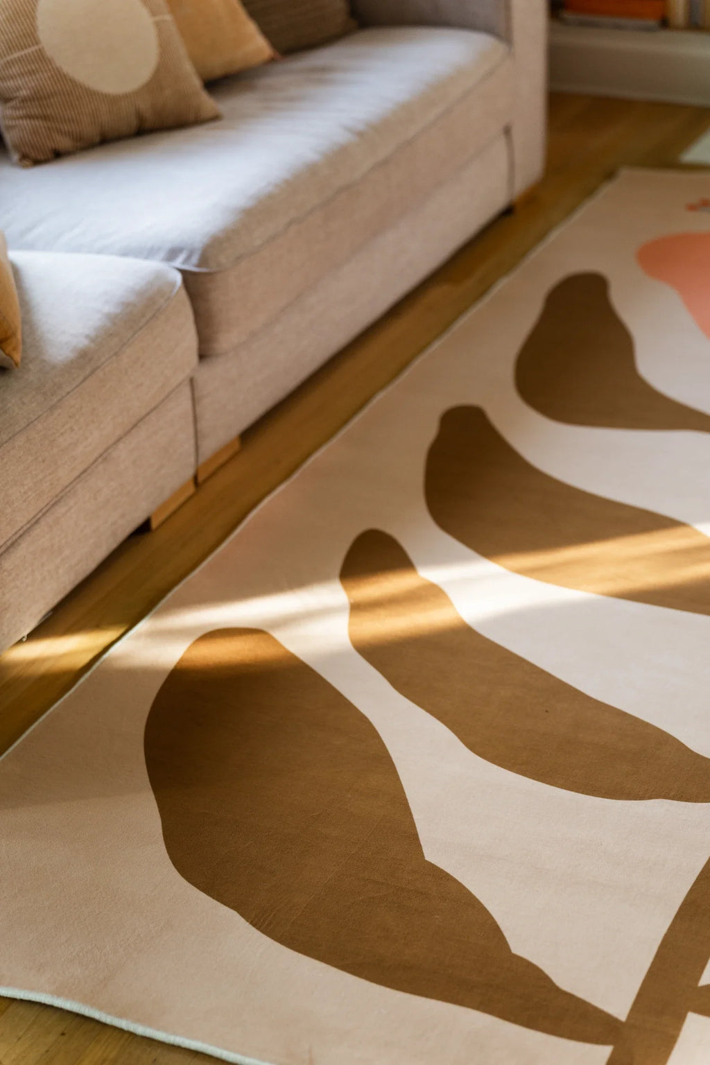 Beige vintage rug beside a neutral sofa with sunlight across the surface and bold botanical shapes on a light wood floor