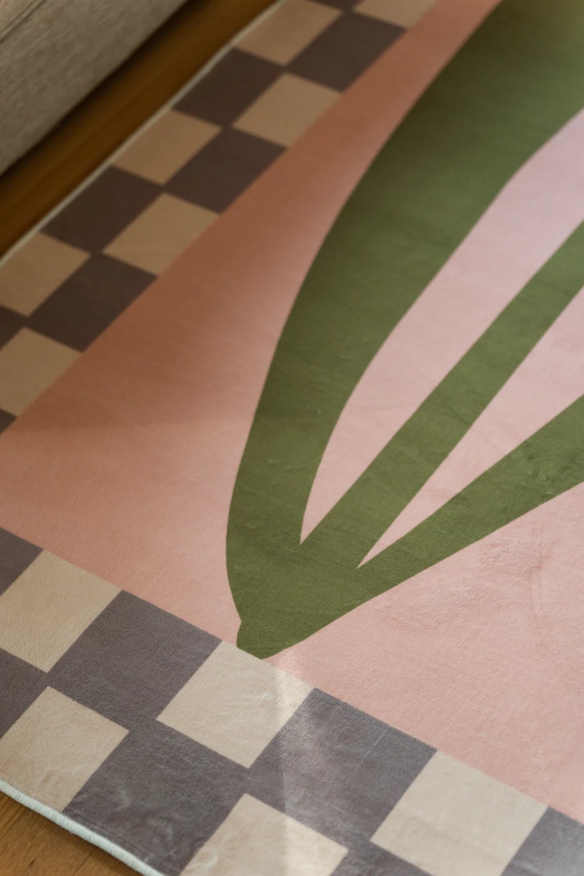 Bottom corner of the blush pink floral rug, showing long olive leaf forms and a soft taupe checkered border