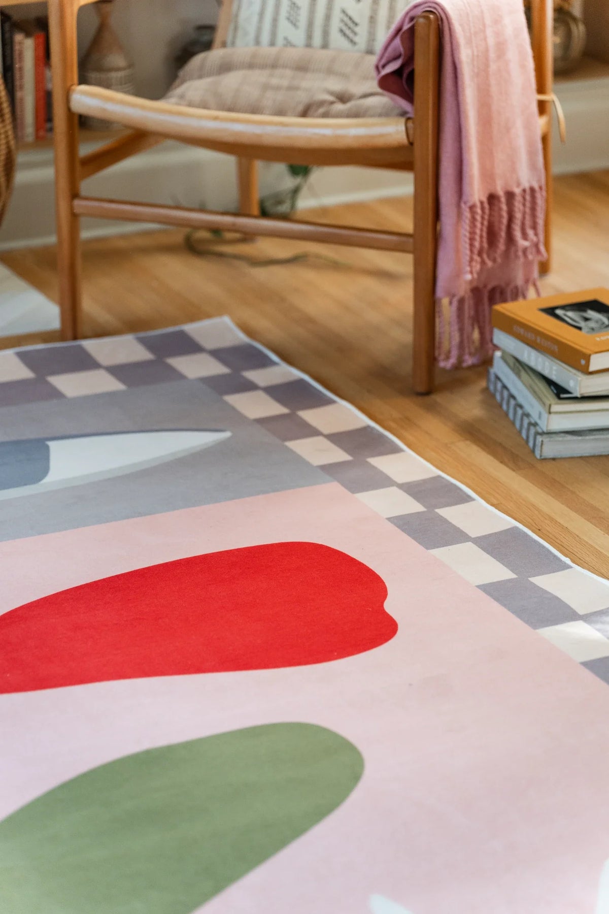 Close crop beside a wood chair and stacked books, highlighting the blush rug's red petal form and soft checkered border