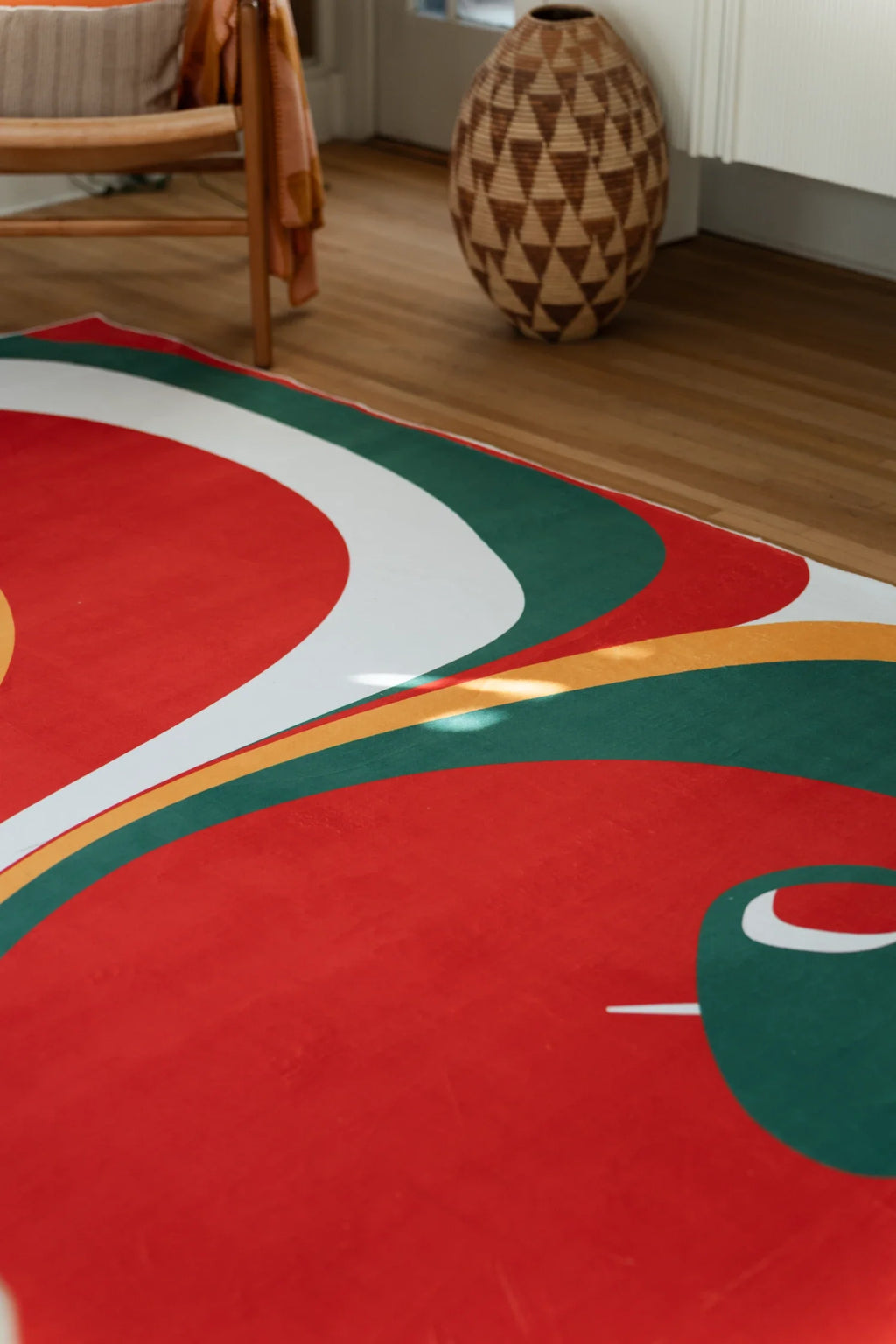 Close up of the olive and red rug beside a chair and woven basket, showing white, green, and red curves