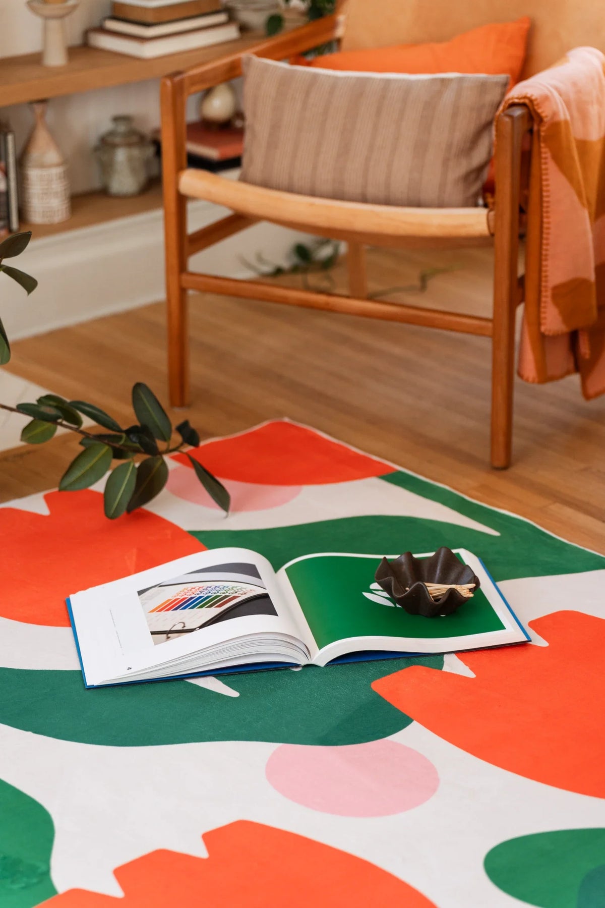 Orange floral area rug near a wood chair and open book, styled on a light wood floor in a relaxed living area