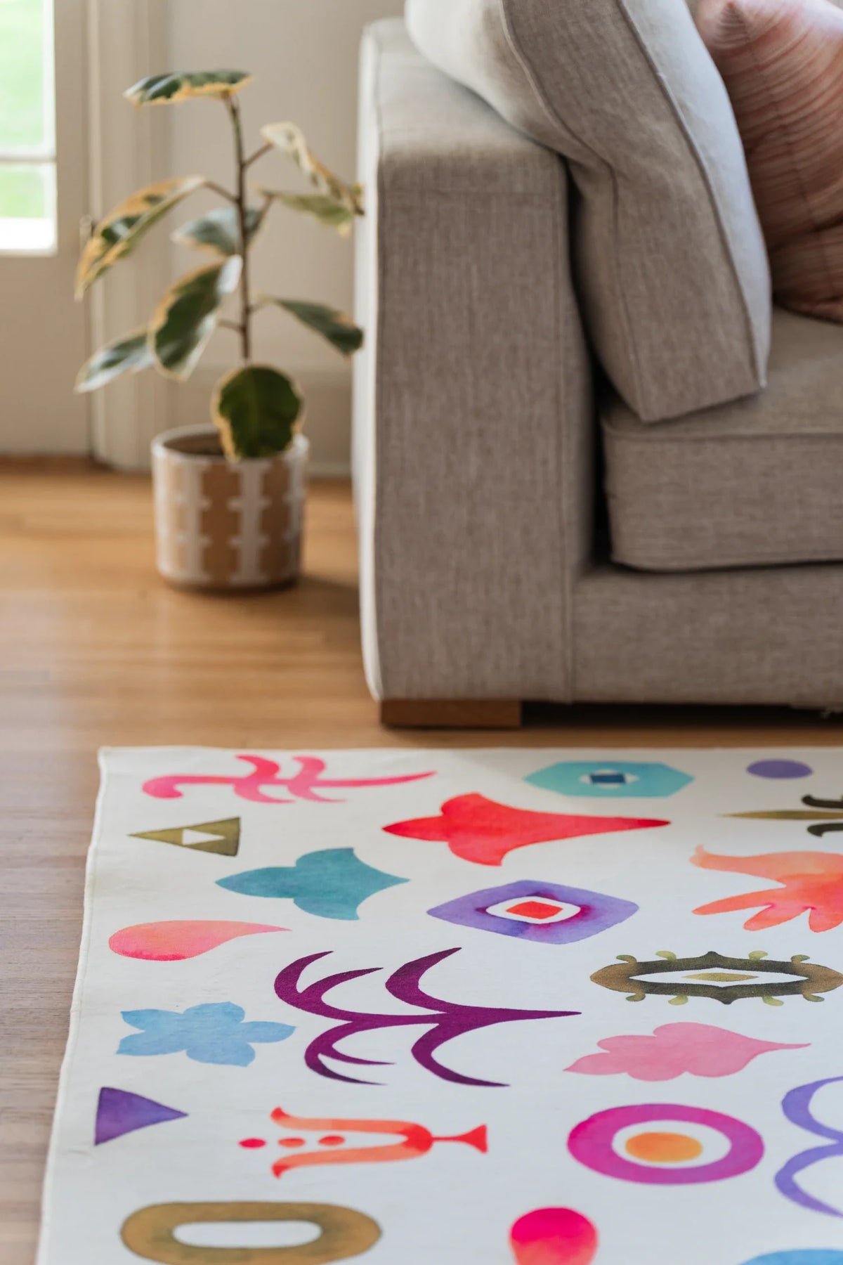 Colorful abstract area rug beside a beige sofa and planter, showing birds, flowers, moons, and small geometric symbols