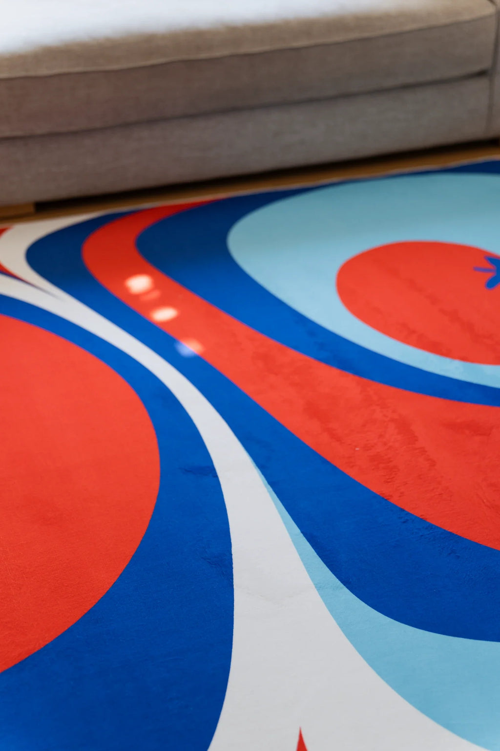 Detail of the Tomato Twist rug with layered red, royal blue, and pale blue bands beside a gray sofa