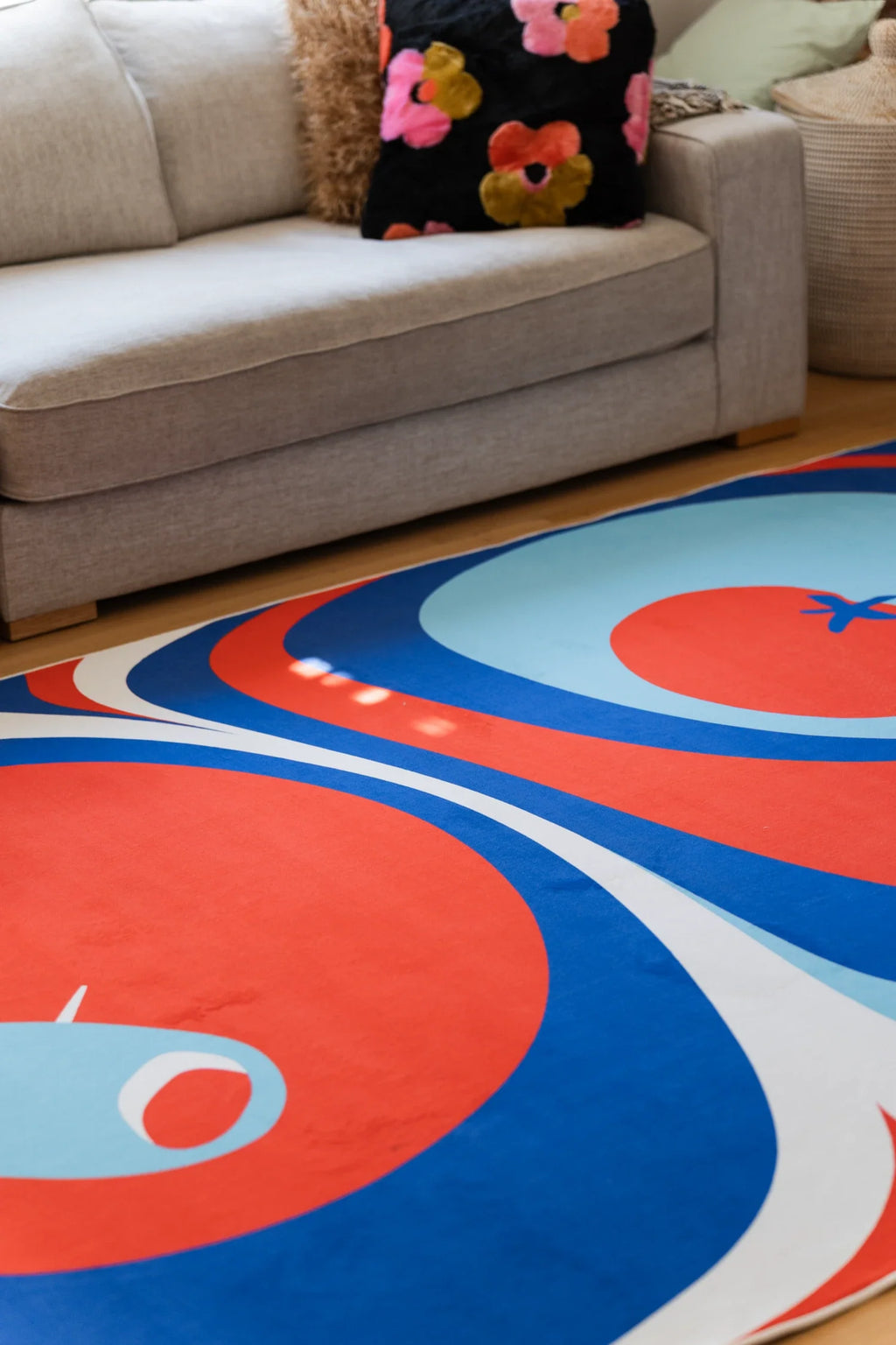 Blue and red abstract rug beside a gray sofa, showing rounded tomato motifs and flowing blue contour bands